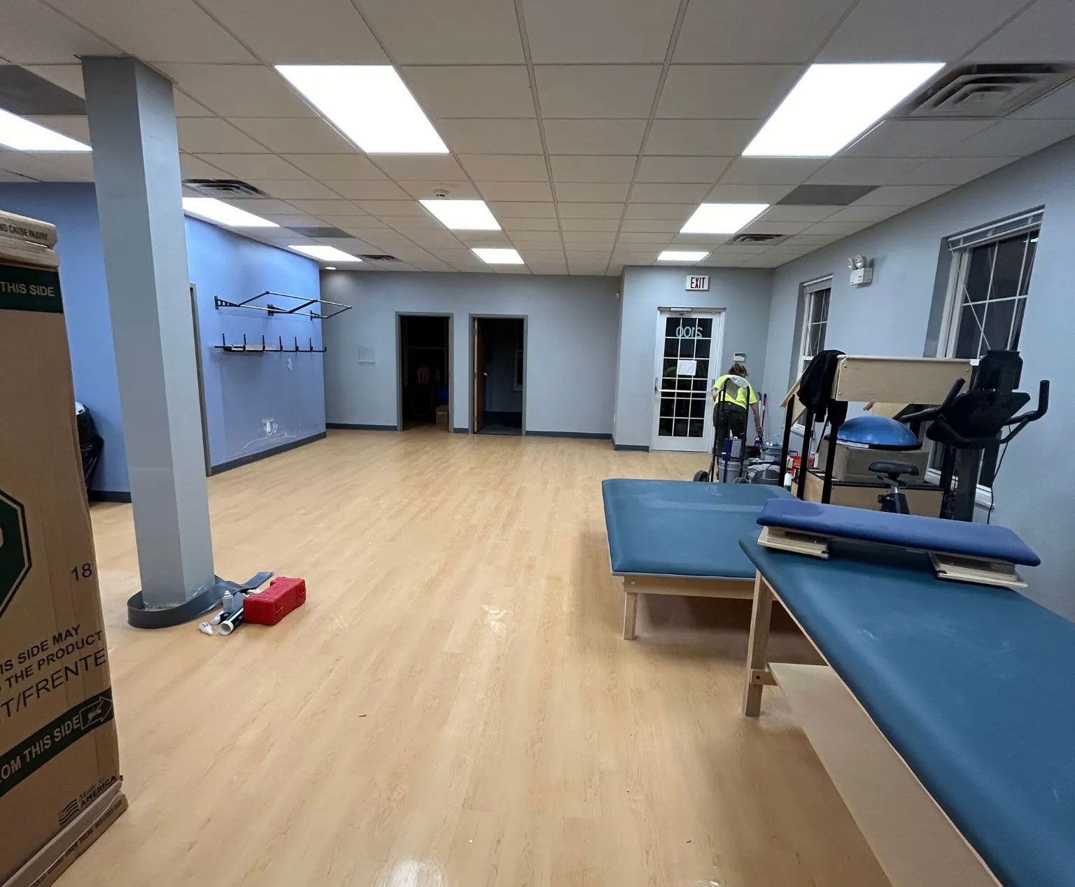 Empty physical therapy room with blue treatment tables, wood floors, and exercise equipment