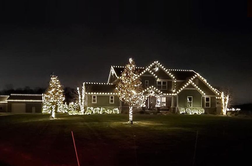 House with Christmas lights at night. Green siding, white lights, and trees are also lit up.