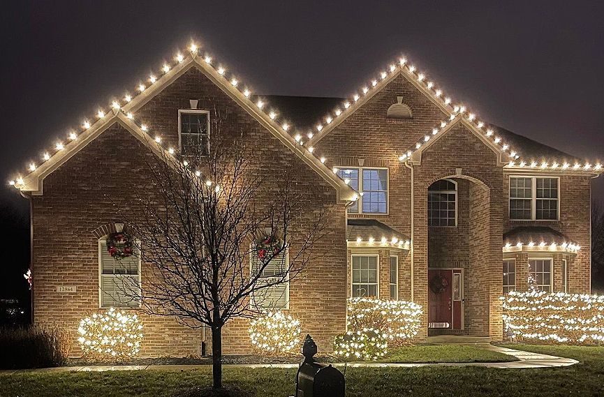 A brick house at night, decorated with white Christmas lights along the roof and bushes.