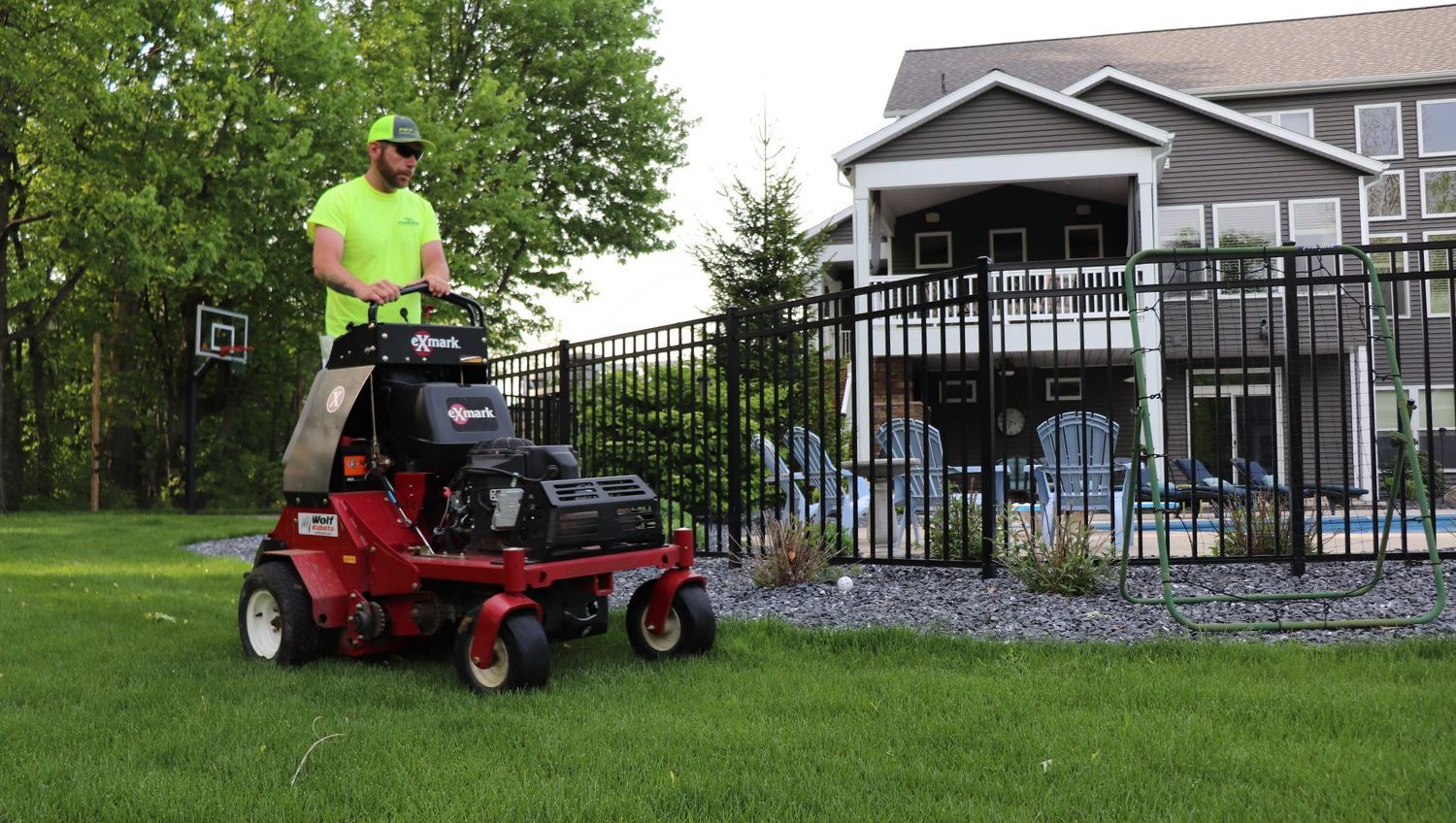 A man mowing a green lawn with a red riding mower near a black fence and a house.