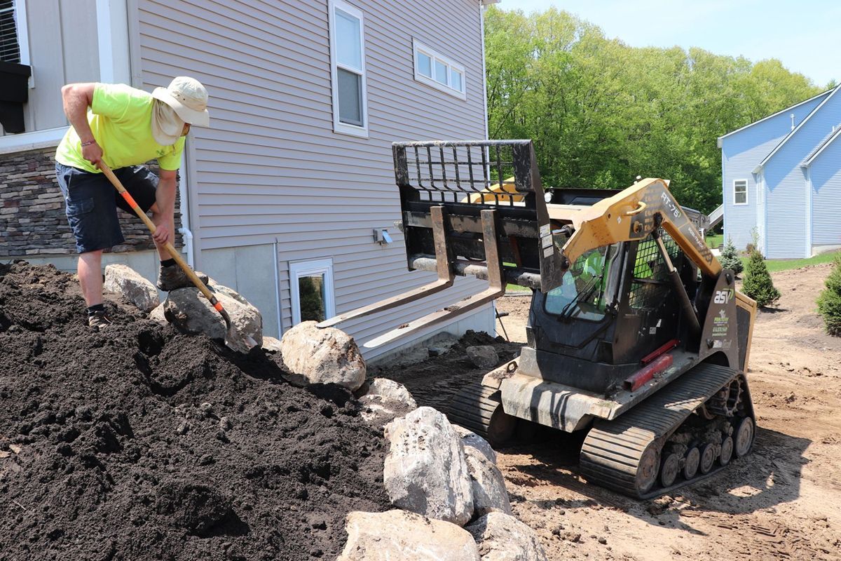 Man shovels rocks alongside a skid steer near a house with tan siding and a blue-roofed building.