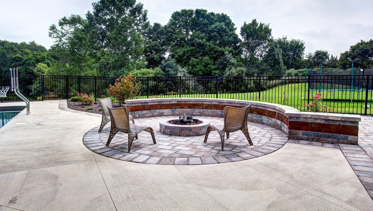 Stone patio with fire pit, chairs, and brick wall, set in a backyard with green trees.