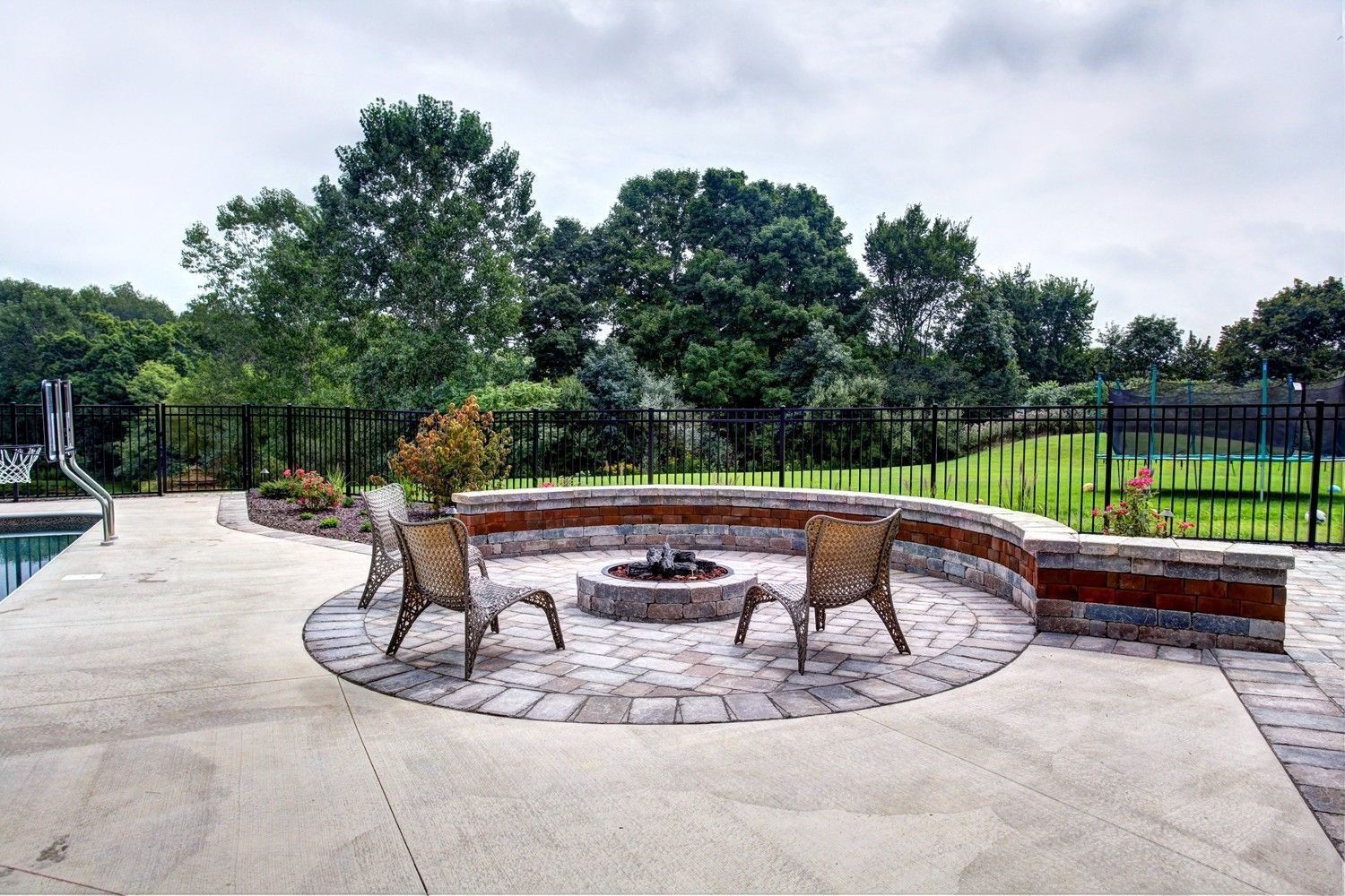 Stone fire pit with chairs on a patio, overlooking a fenced yard with trees.