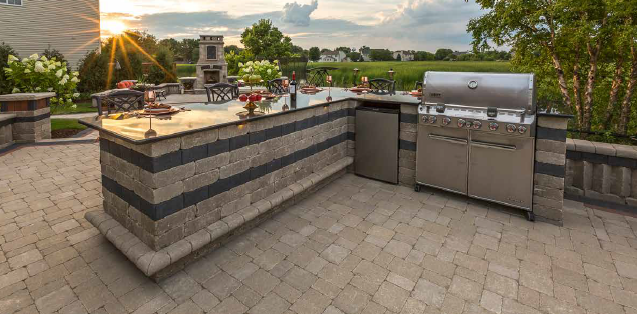 Outdoor kitchen with a built-in grill, bar, and refrigerator on a stone patio with a sunset in the background.