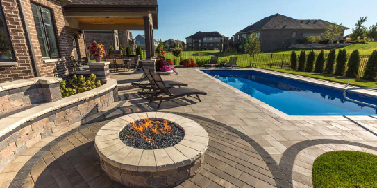 Backyard with a pool, patio, fire pit, and a house in the background on a sunny day.