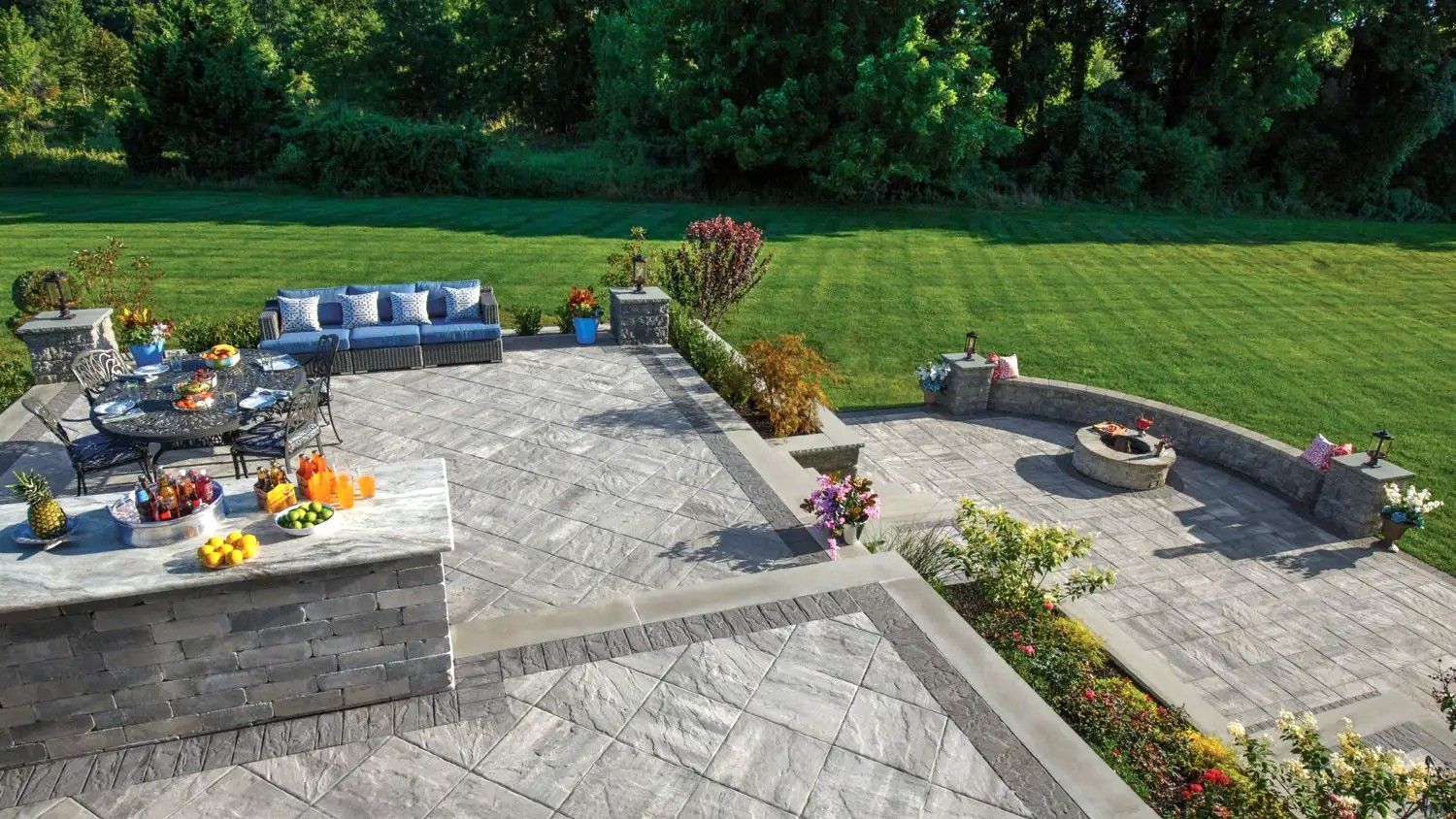 A landscaped backyard with patio, seating areas, fire pit, and lawn, surrounded by green trees and a sunny sky.