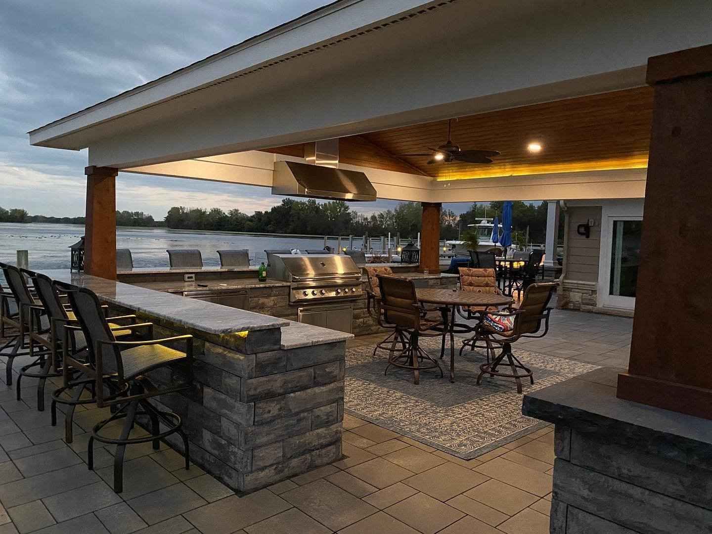 Outdoor kitchen with bar, grill, seating, and water view under a covered patio with lights.
