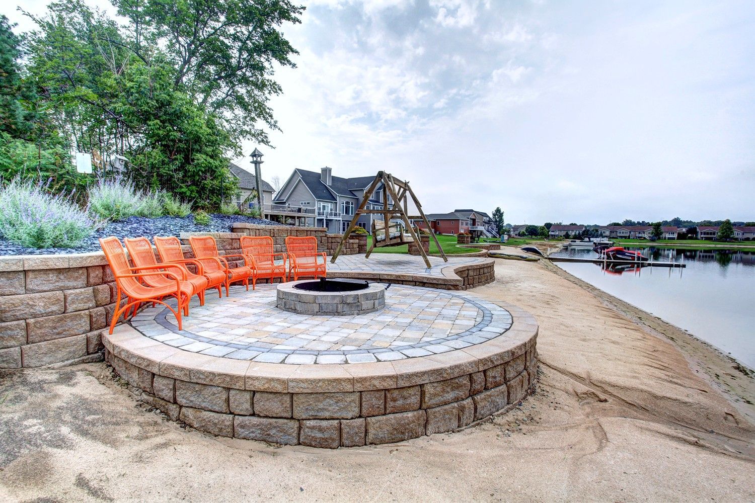 A fire pit is surrounded by chairs on a patio next to a lake.