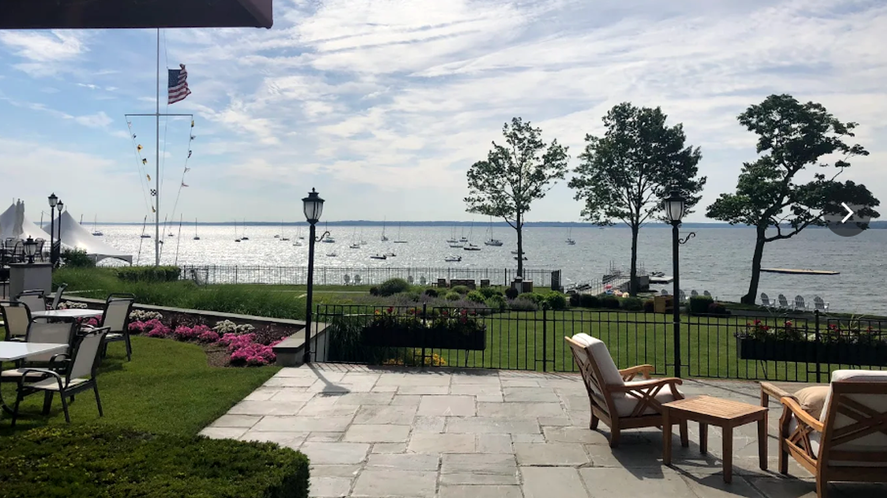 Patio seating and lawn overlooking a calm waterfront with trees, lamps, and an American flag