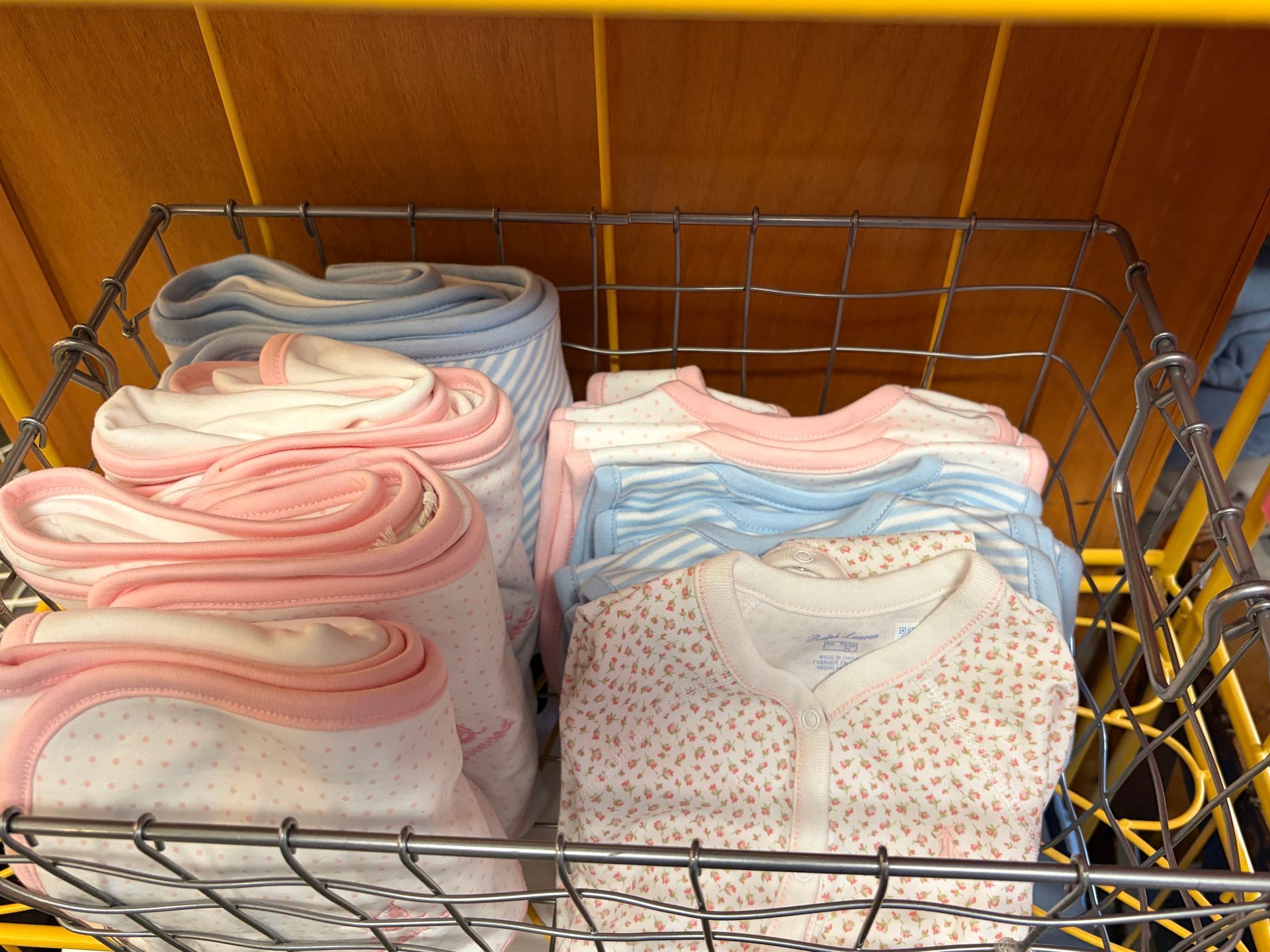 Folded pastel baby clothes in a wire basket on a wooden shelf.