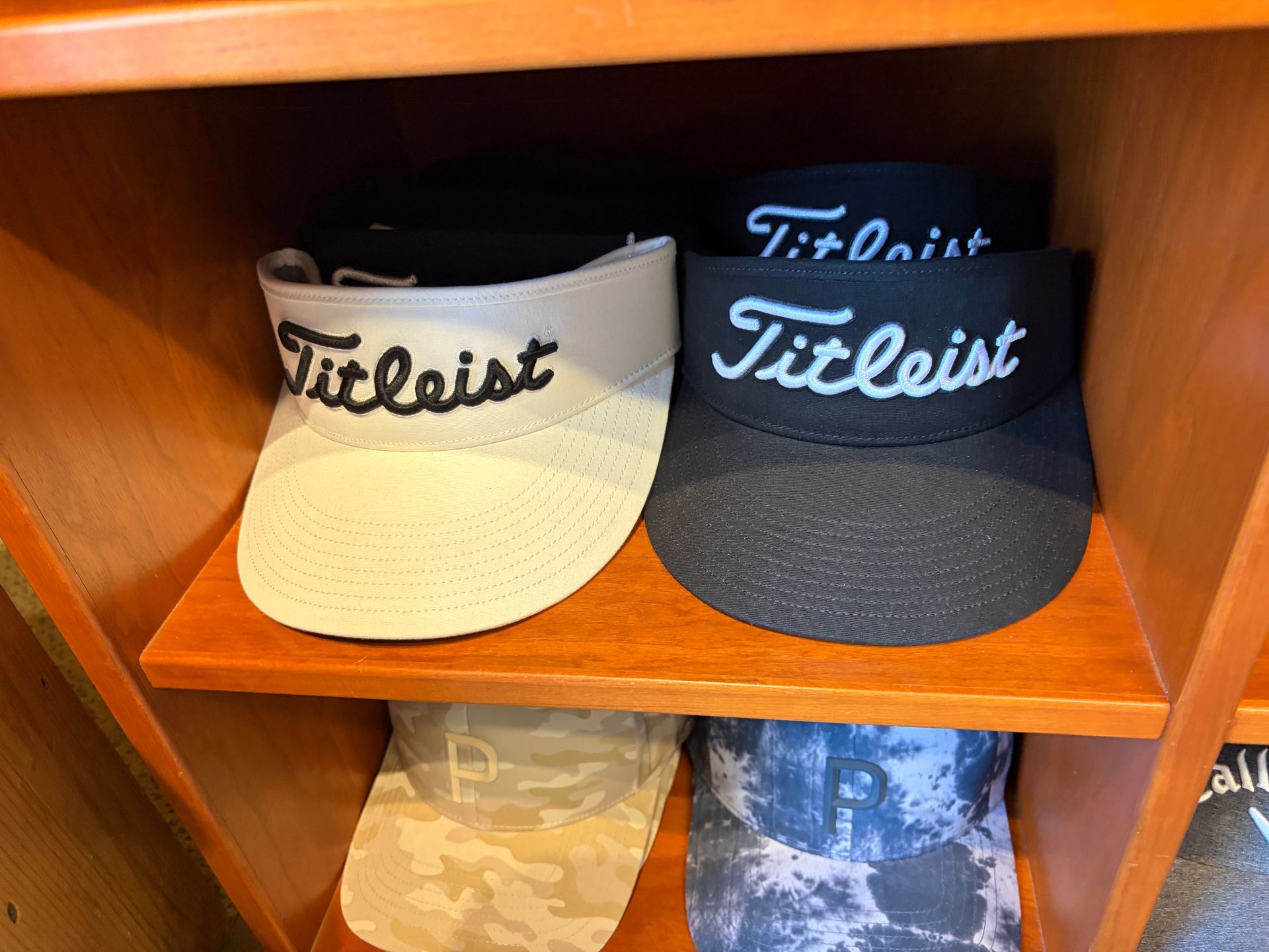 Two Titleist visors on a wooden shelf, one cream and one black, with camo hats below