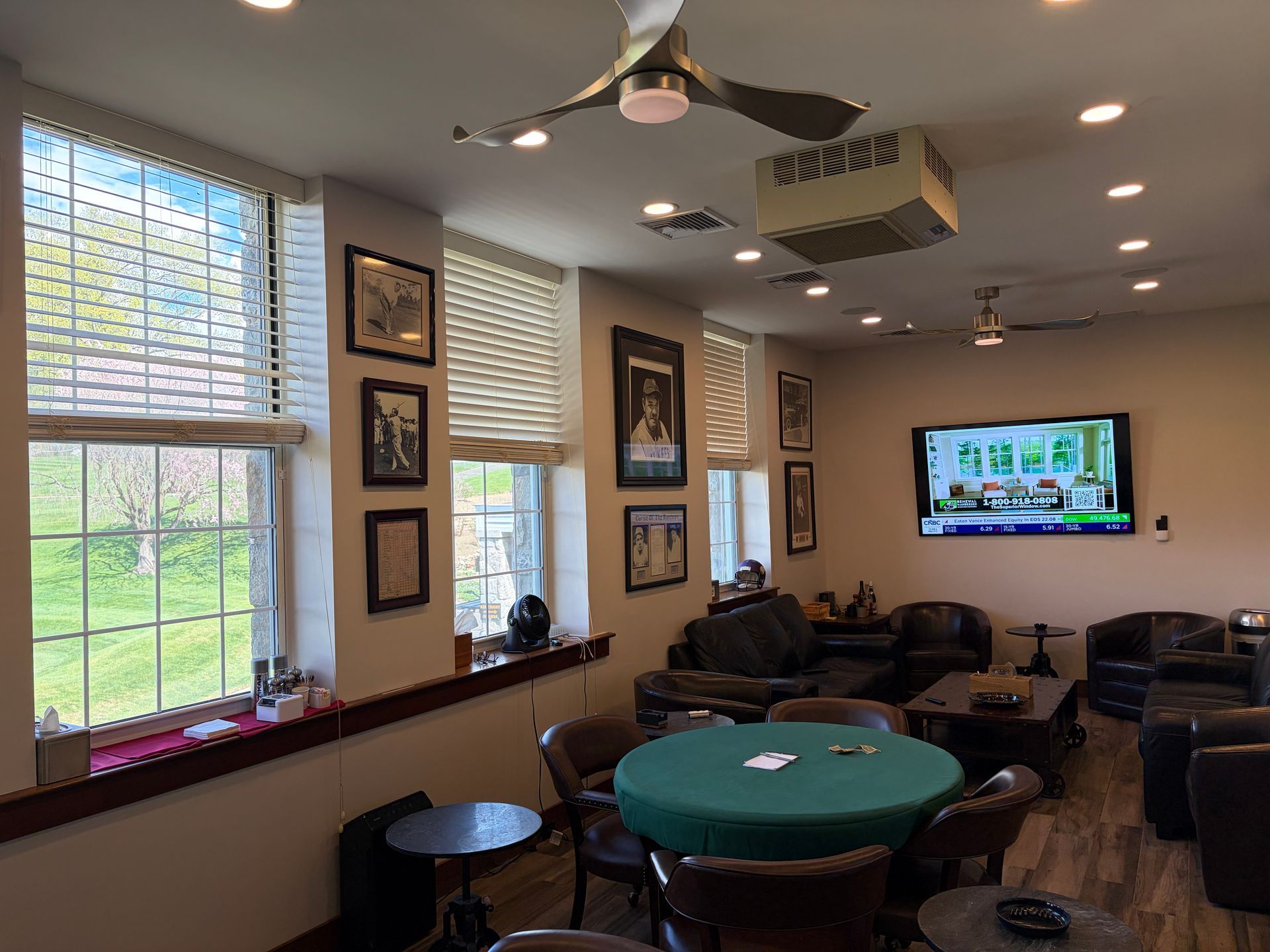 Lounge with windows, ceiling fans, framed art, and a wall-mounted TV above a green poker table.