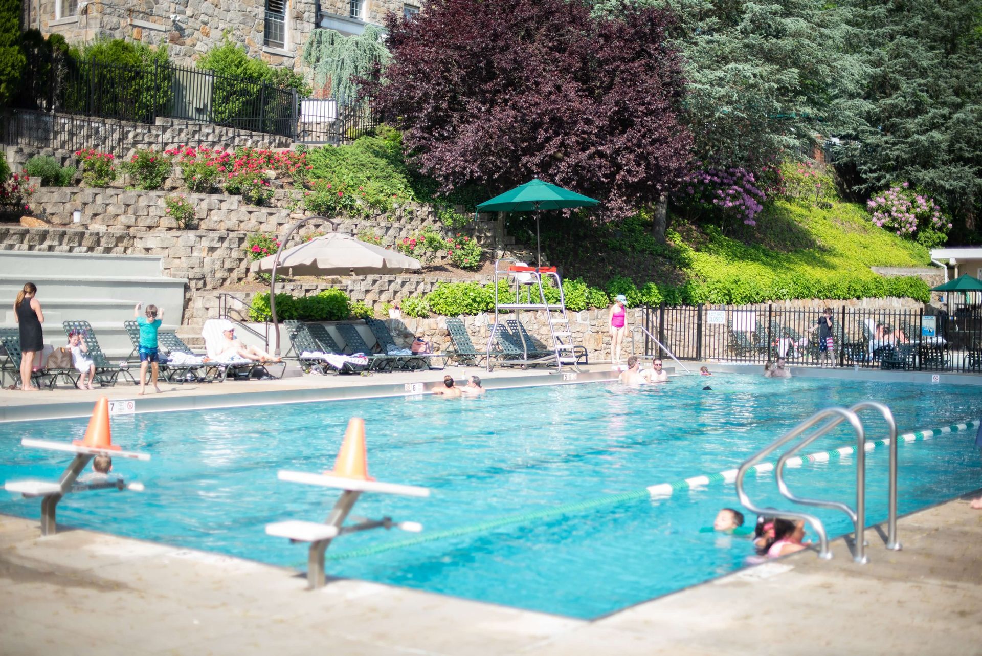 Outdoor swimming pool with people, surrounded by lush green hillside and stone buildings.