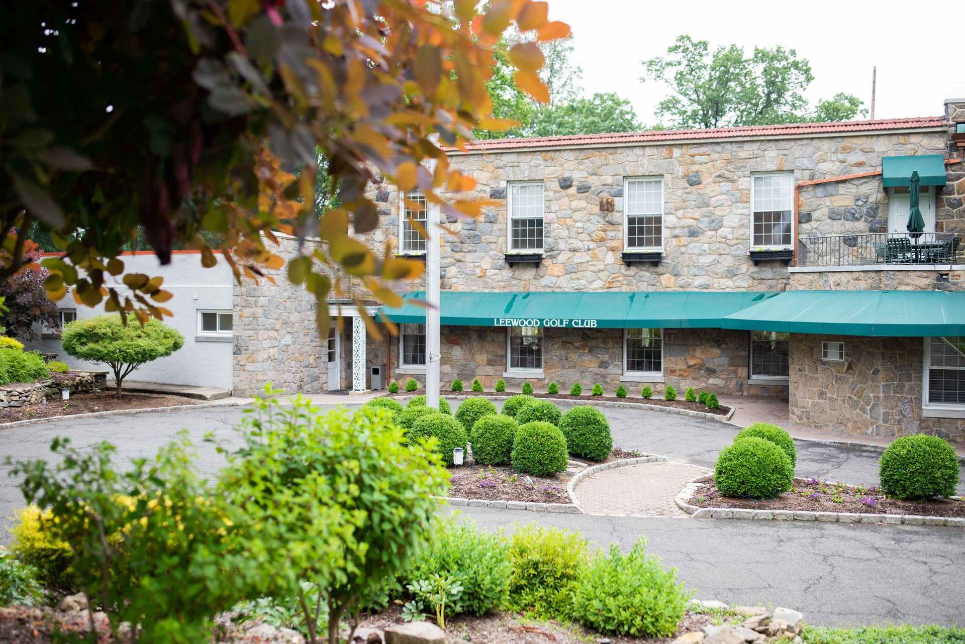 Courtyard with stone building, teal awnings, and landscaped green shrubs under overhanging autumn leaves