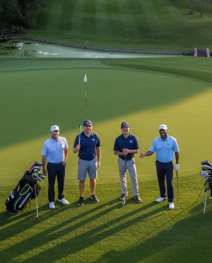 Four golfers standing on a green, holding clubs. Golf bags beside them. Setting appears to be a golf course at dusk.