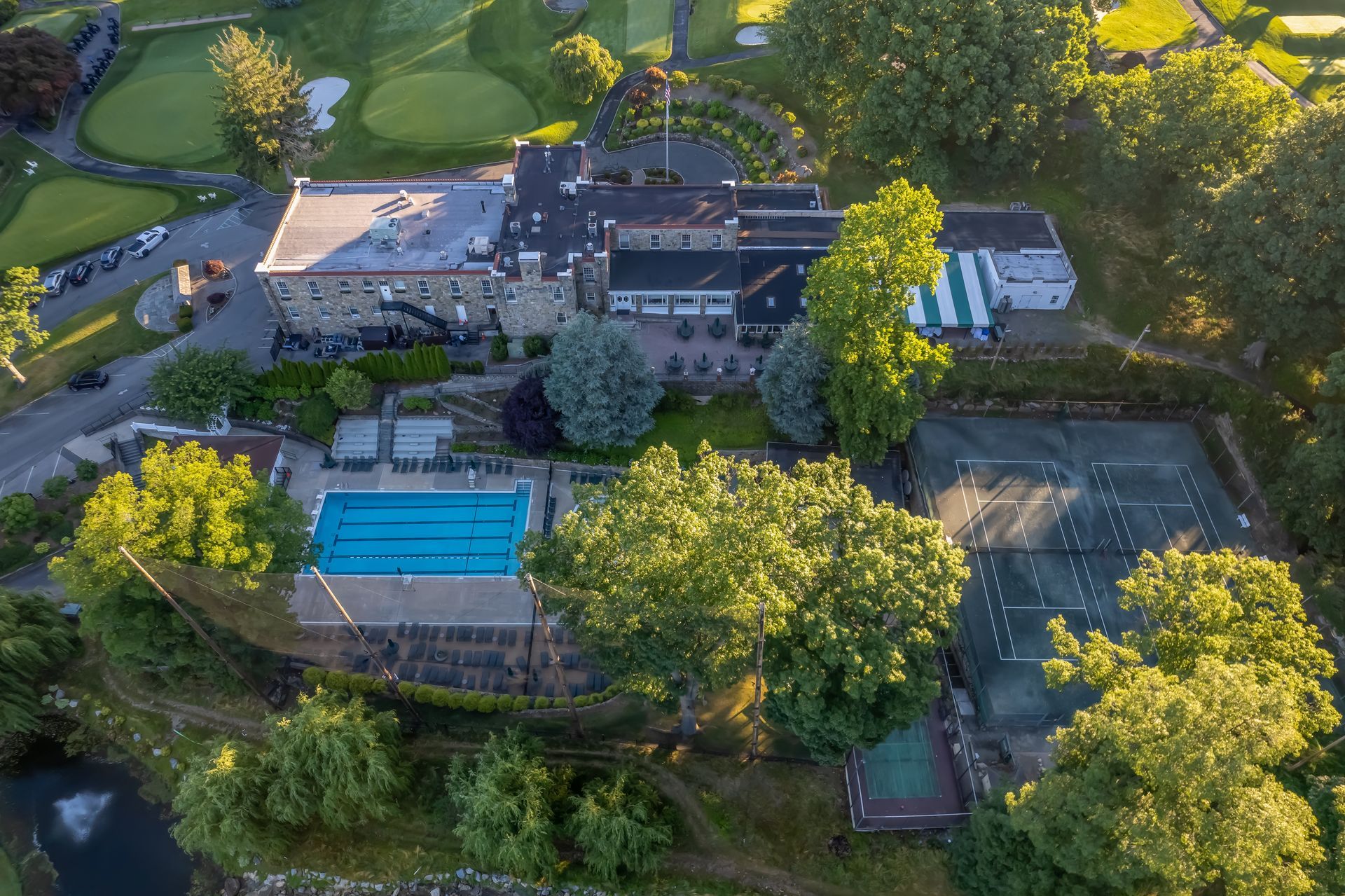 Aerial view of a country club with a building, swimming pool, tennis courts, and golf course surrounded by trees.