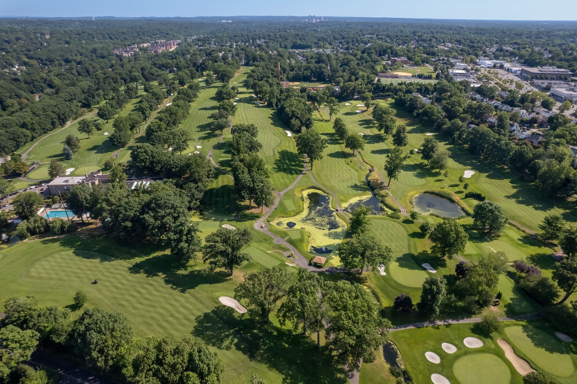 Aerial view of a green golf course with trees, fairways, and water hazards on a sunny day.