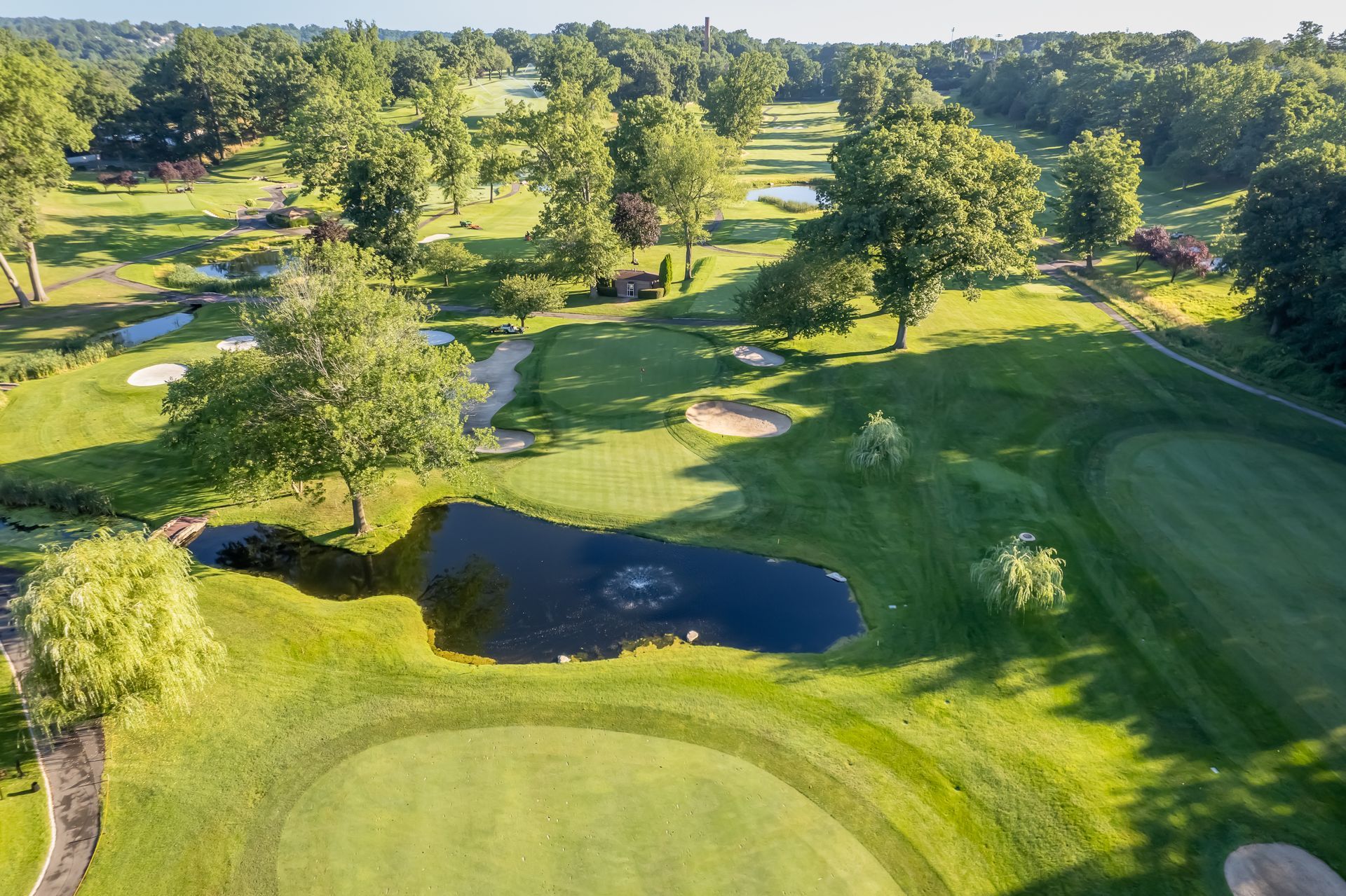 Aerial view of a green golf course with a pond, sand traps, and trees under a bright sky.