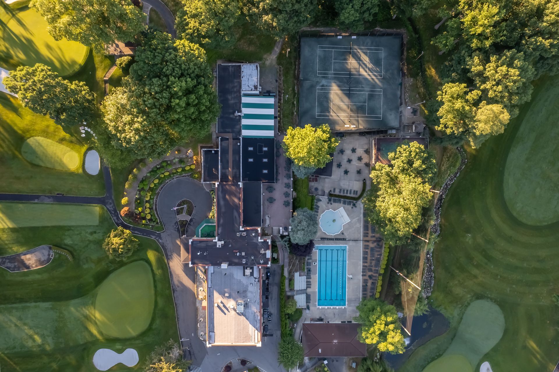 Aerial view of a clubhouse with a tennis court, pool, and golf course. Lush greenery surrounds the structures.