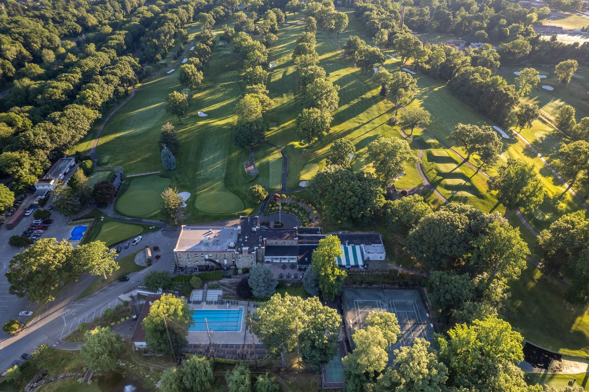 Aerial view of a golf course with a clubhouse, pool, tennis courts, and surrounding trees.