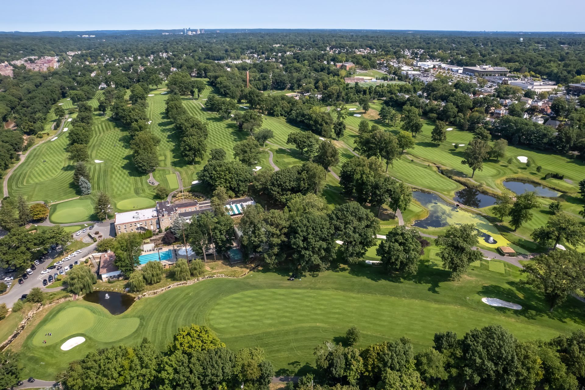 Aerial view of a lush green golf course with trees, ponds, and buildings. Sunny day with blue sky.