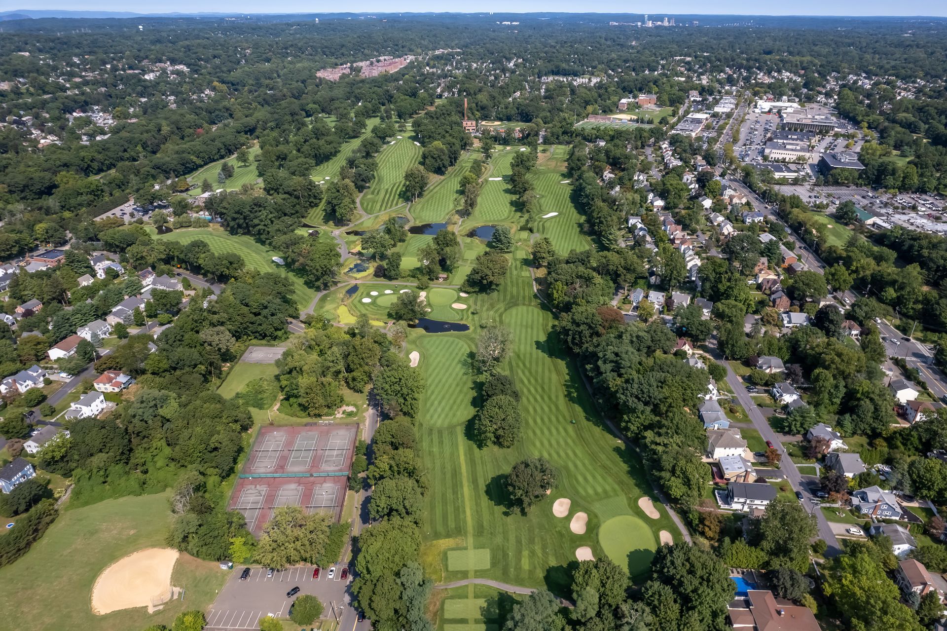 Aerial view of a green golf course surrounded by trees and residential houses under a blue sky.