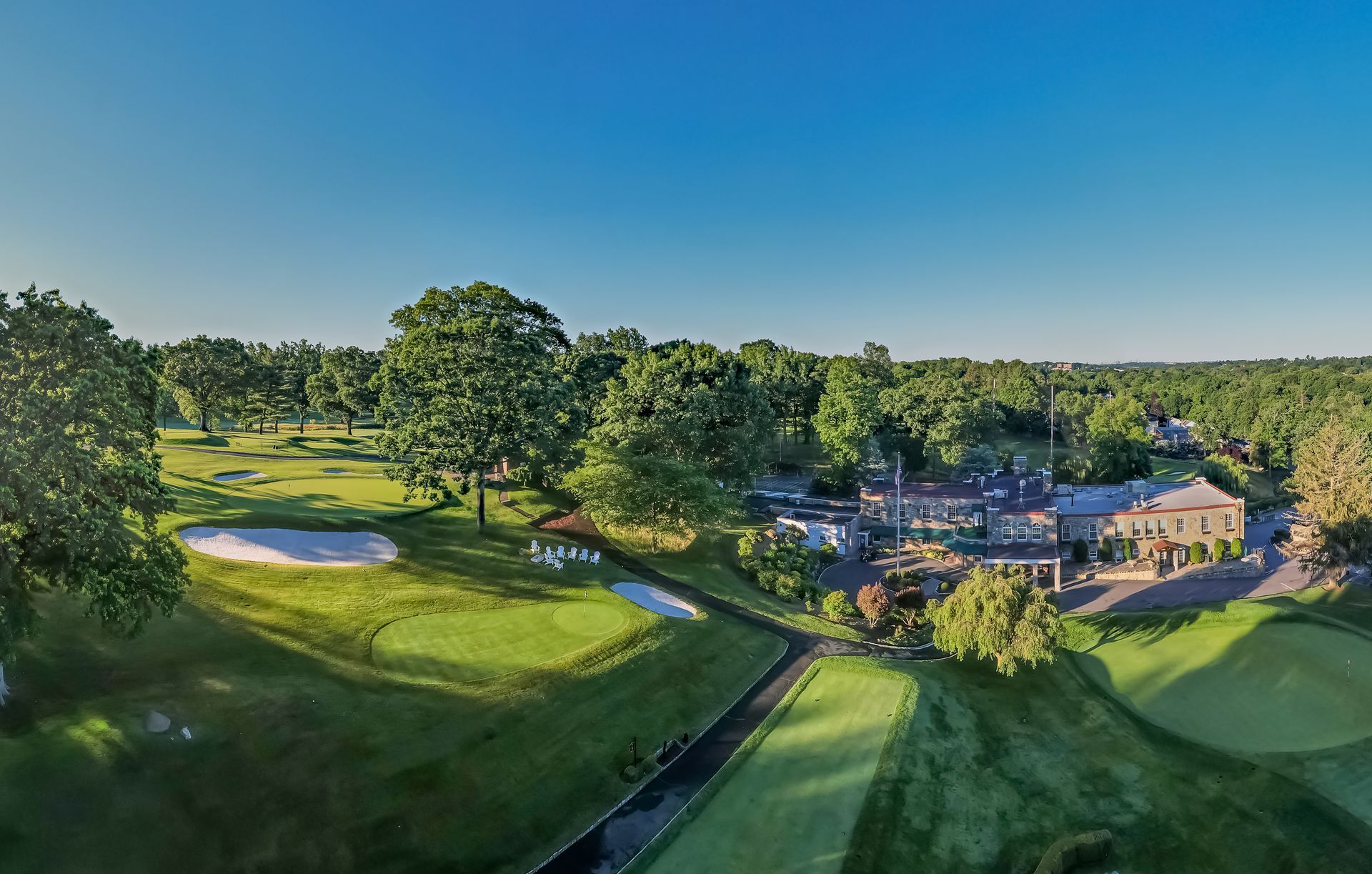 Aerial view of a golf course with green fairways, a sand trap, and a large building surrounded by trees.