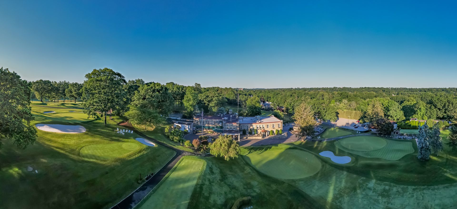 Aerial view of a green golf course with a clubhouse, trees, and a clear blue sky.