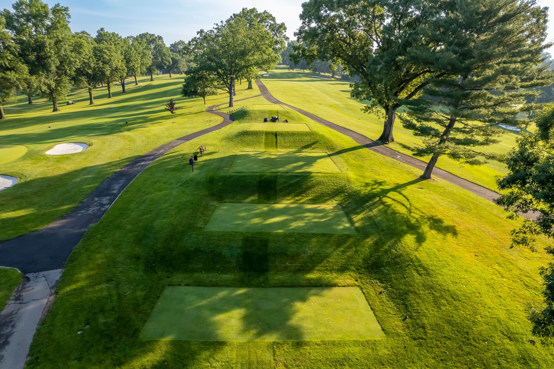 Green golf course with tiered tee boxes, trees, and fairways in sunlight.