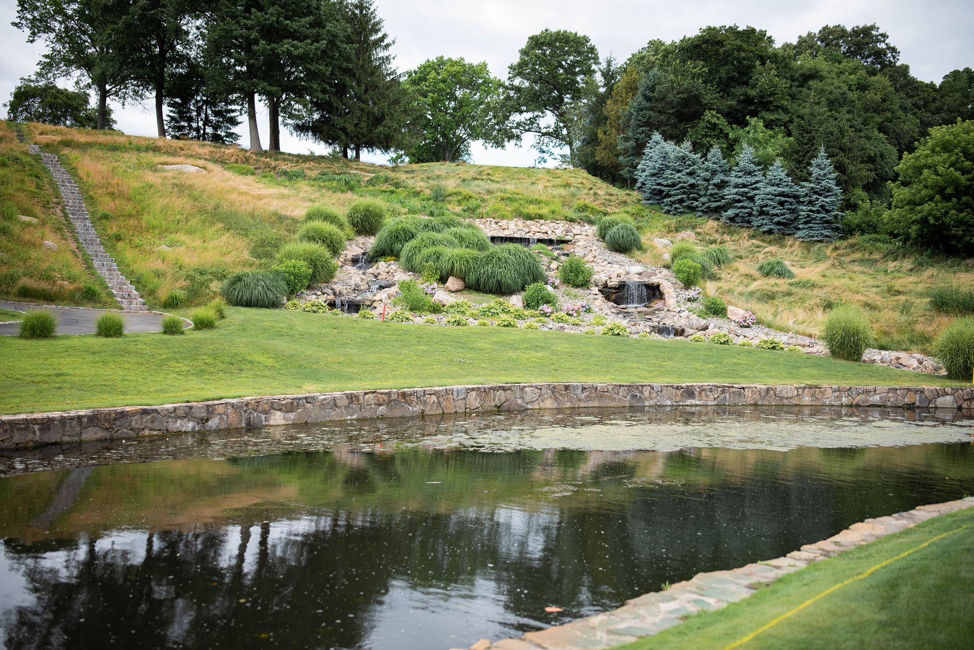 Pond with stone wall, small waterfall on grassy hill, trees in the background, overcast sky.