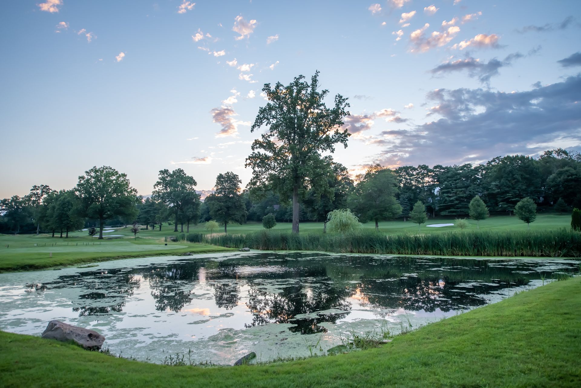Pond on a golf course under a cloudy sky. Lush green grass surrounds the water, with trees in the distance.