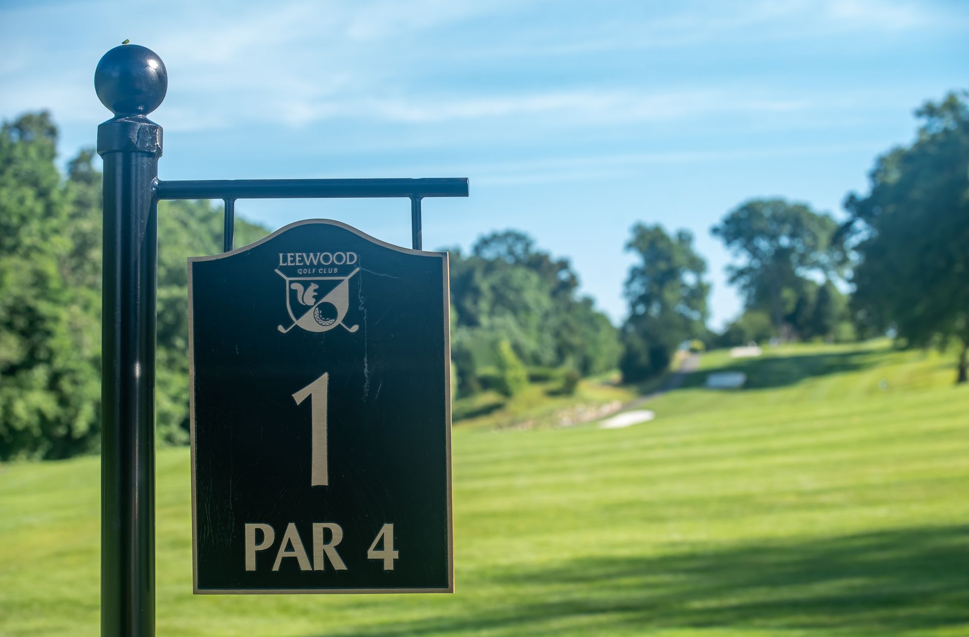 Golf course sign for hole 1, a par 4. Green grass, blue sky, trees in the background.