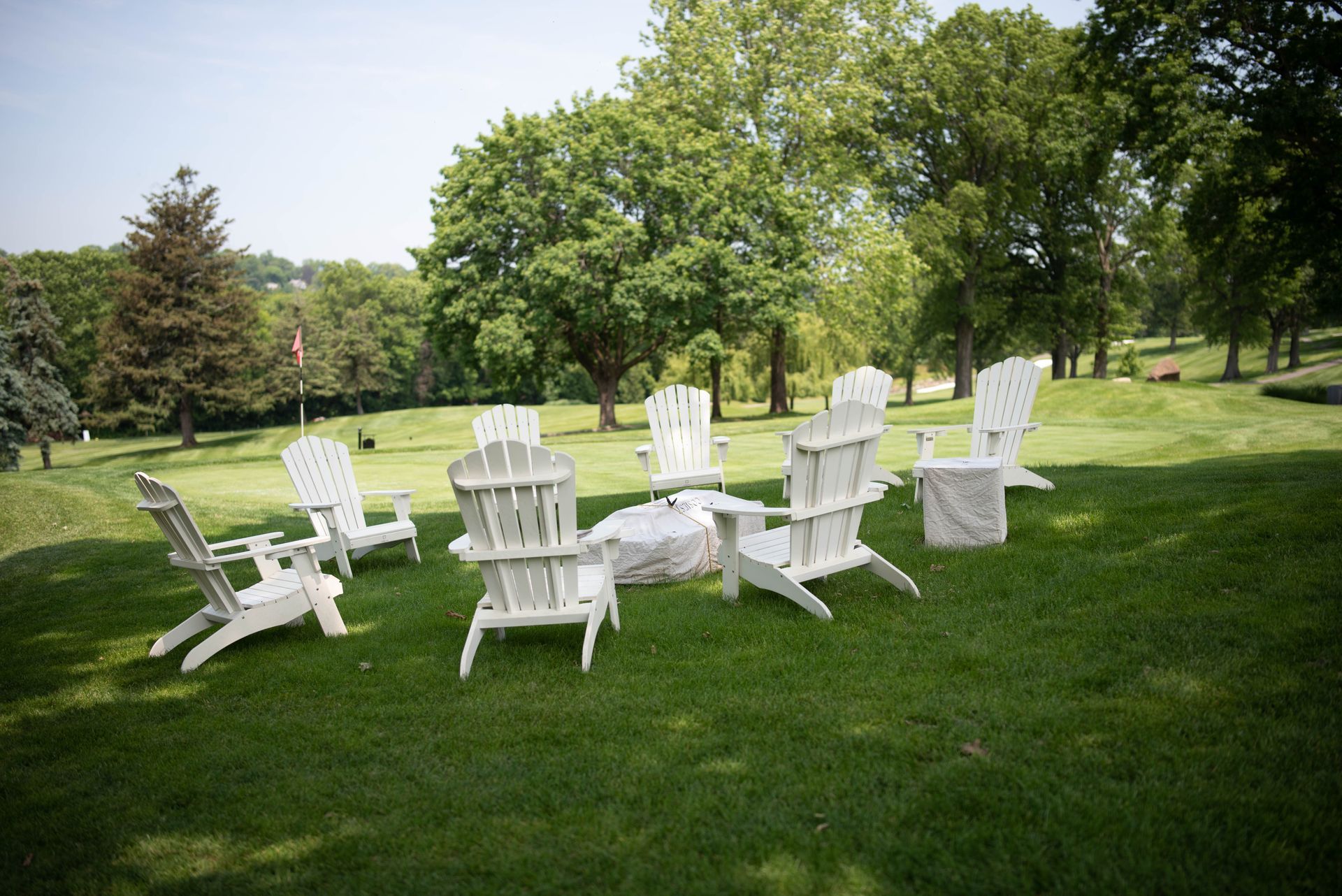 White Adirondack chairs circle a fire pit on a grassy lawn, trees in the background.