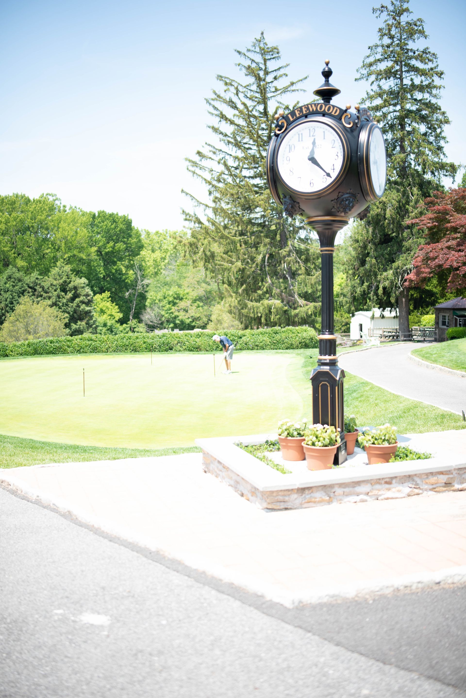 Black clock tower with white clock face, set on a stone base, near a golf course green.