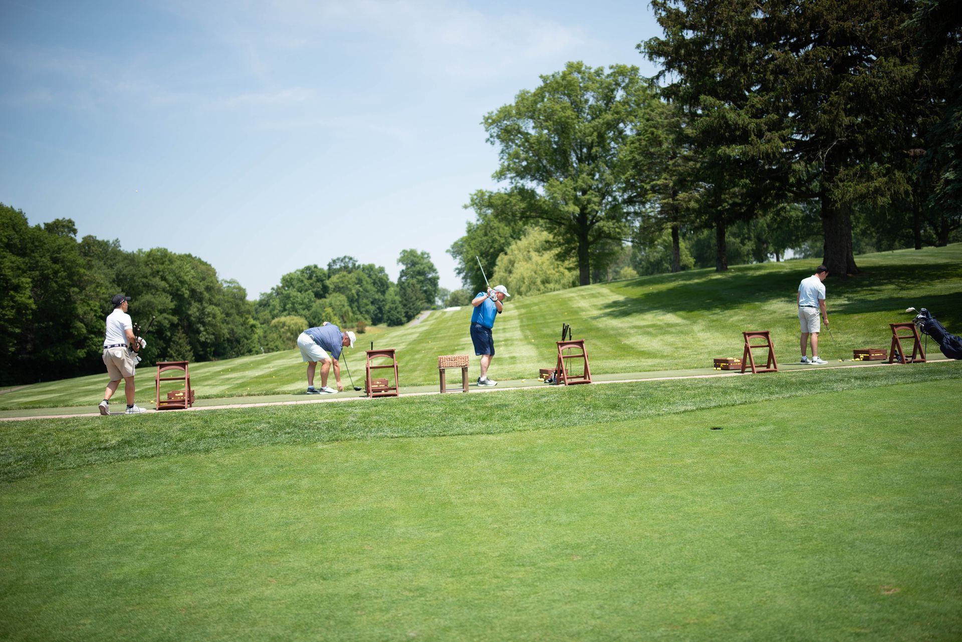 People practicing golf swings on a green, sunny day. Wooden stands hold golf balls, trees in the background.