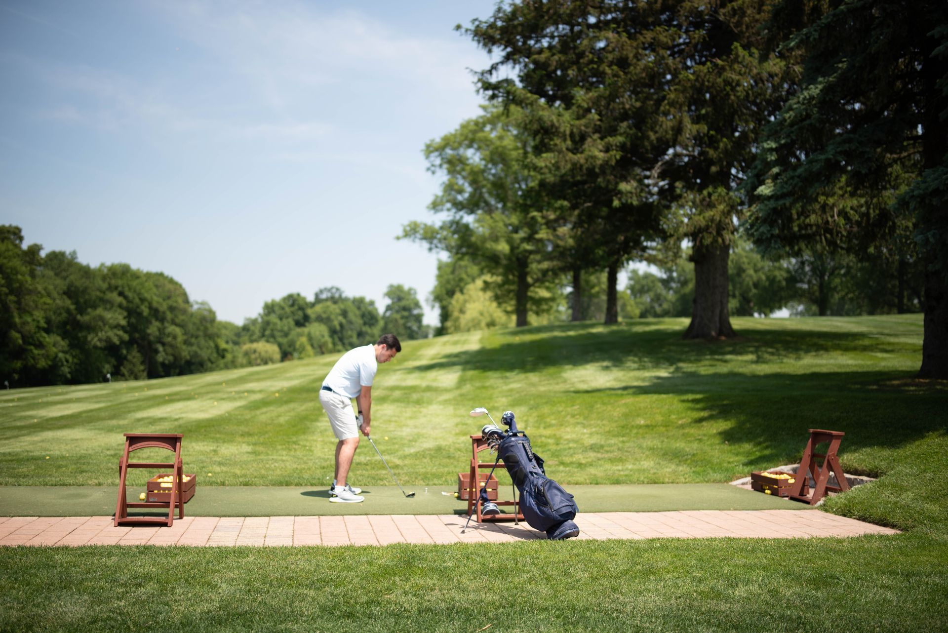 Person on a golf course about to swing. Green grass, blue sky, trees in the background. Golf bag and equipment nearby.