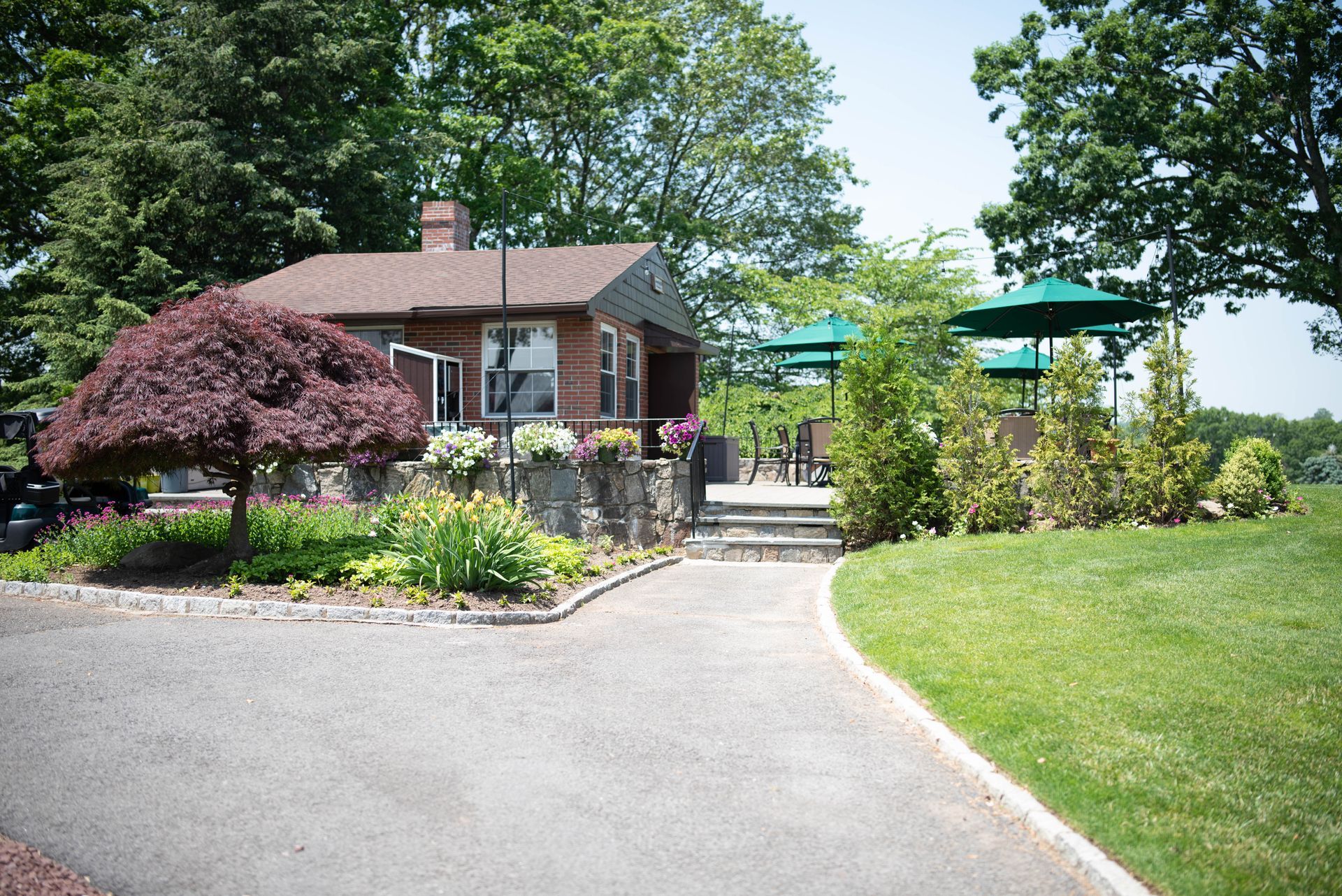 Brick building with stone wall and driveway, surrounded by greenery and trees.