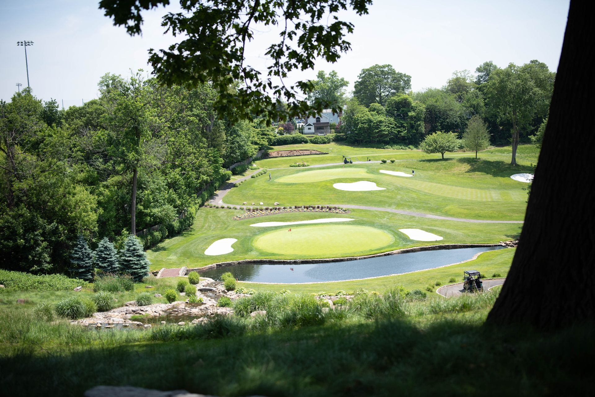 Golf course with green fairways, sand traps, and a pond surrounded by trees on a sunny day.