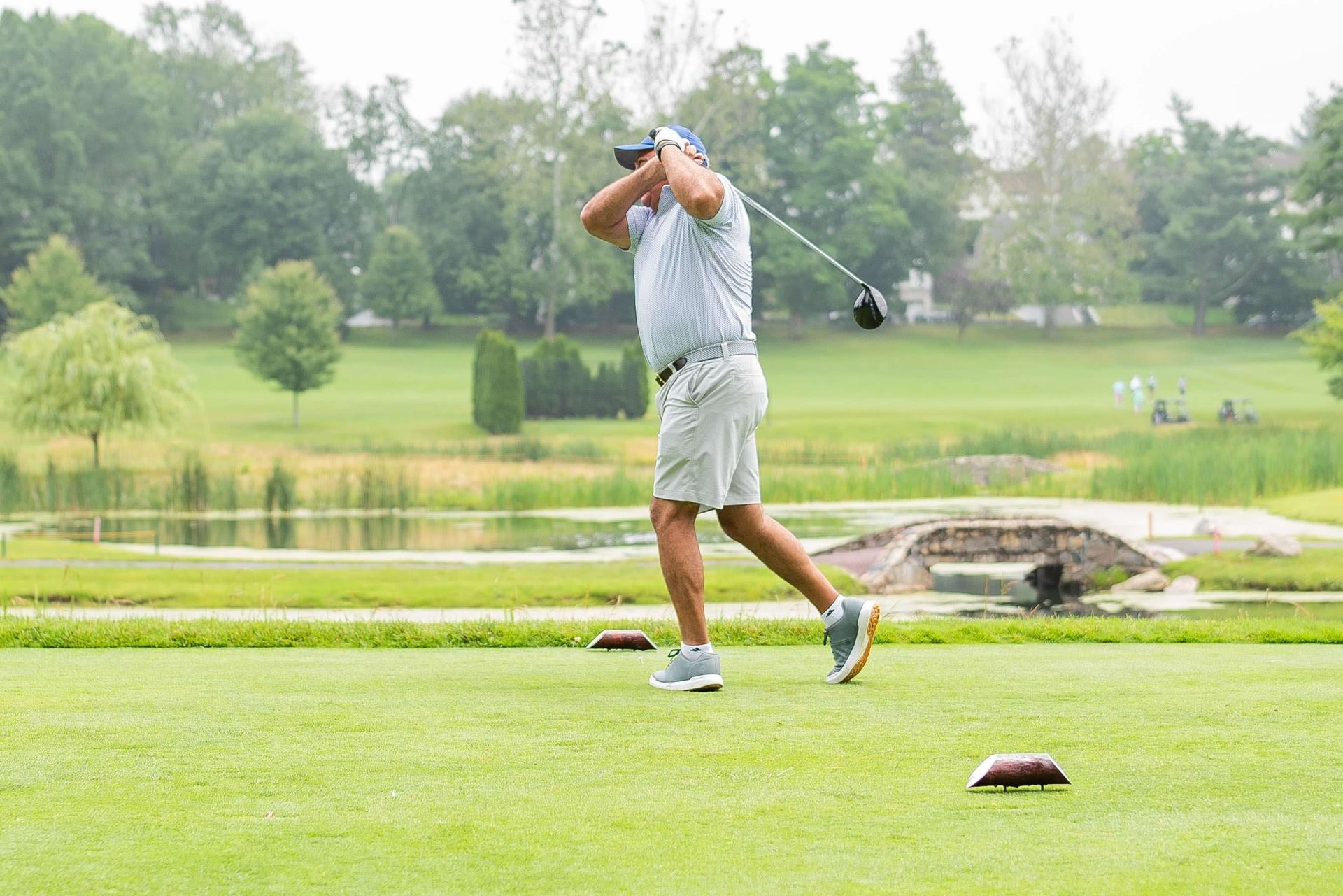 Golfer swinging a club on a green course, trees and water in the background.