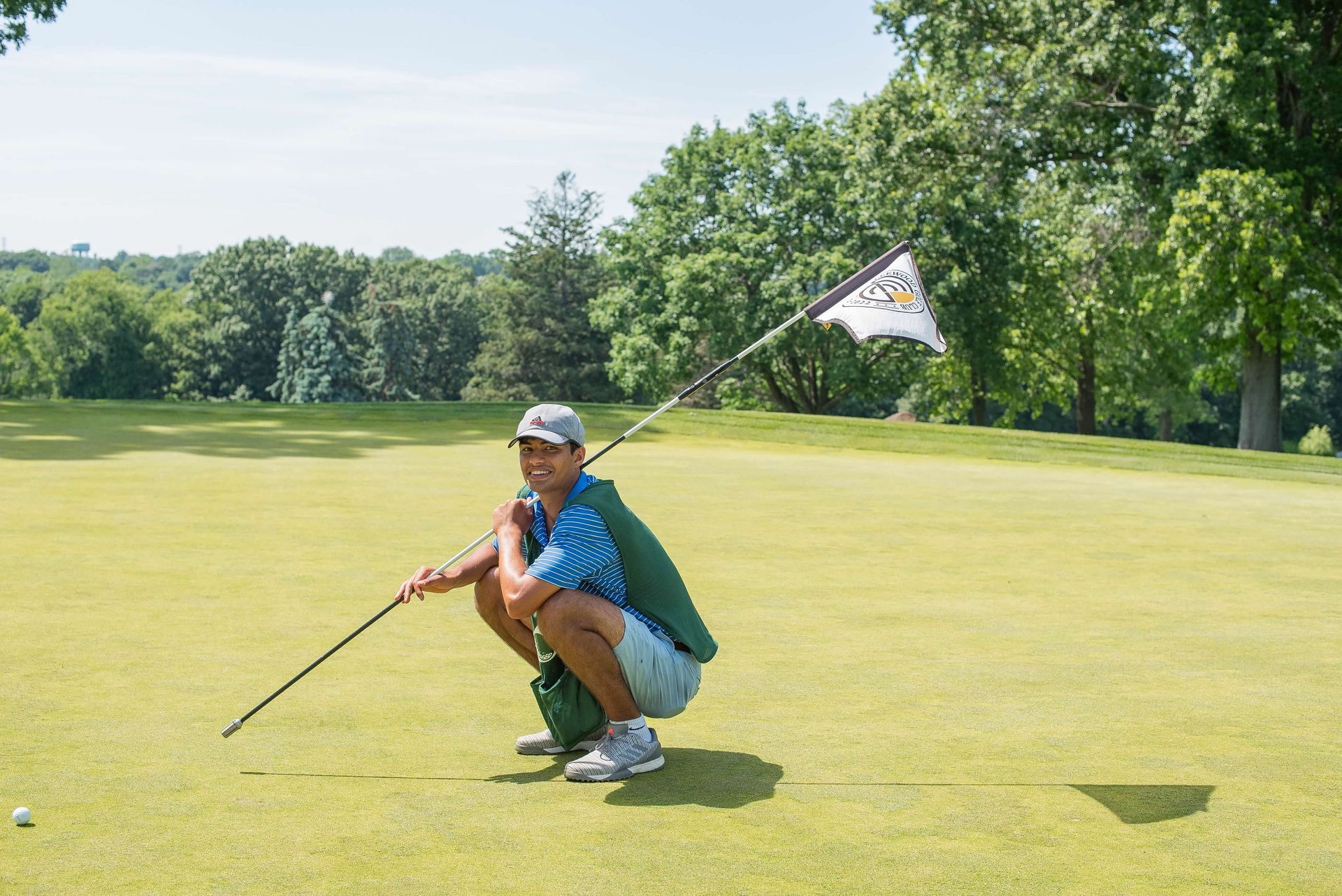 Golfer squats on green, flag in hand. Sunny day, trees in background, golf ball on grass.