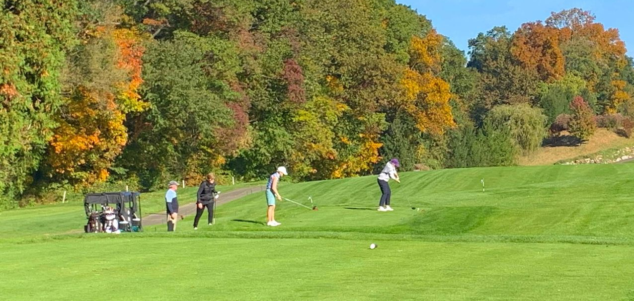 Golfers on a green fairway beside autumn trees under a clear blue sky