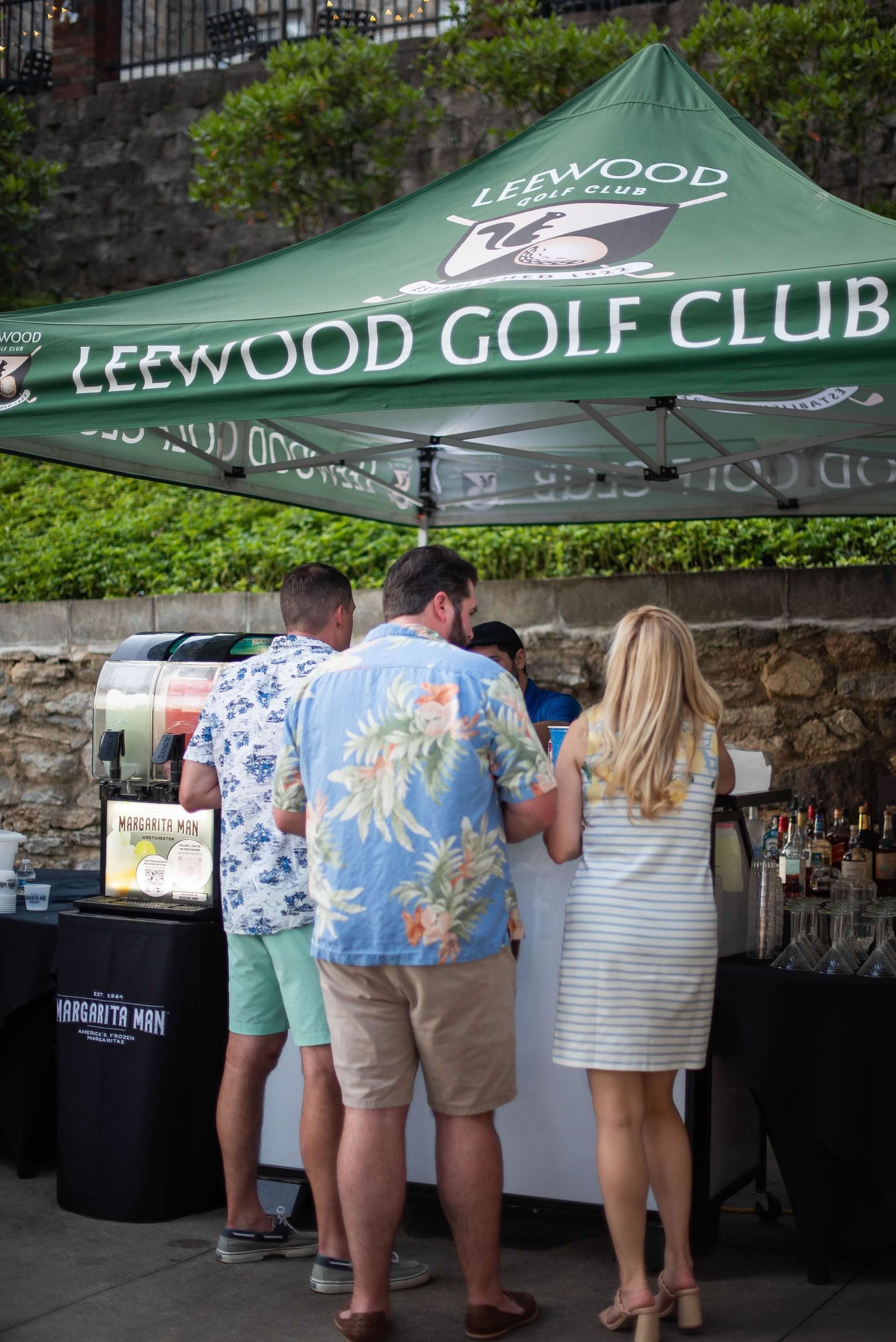 People standing at a Leewood Golf Club outdoor beverage stand under a green canopy.