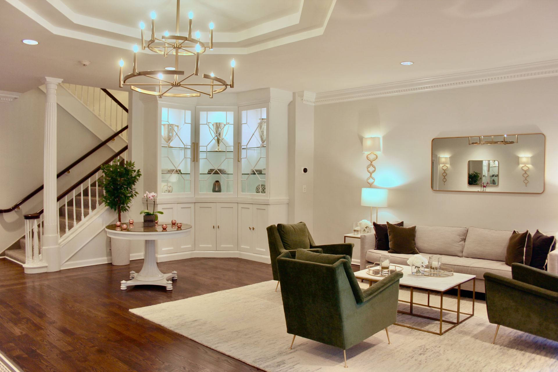 Elegant living room with green chairs, white sofa, staircase, hardwood floors, and chandelier lighting