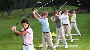 Four youth are practicing their golf swings on a green course, holding clubs mid-swing while wearing golf attire.