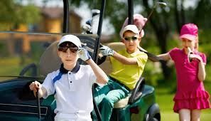 Three children in golf attire pose around a golf cart on a green course; one sits inside while two stand nearby.