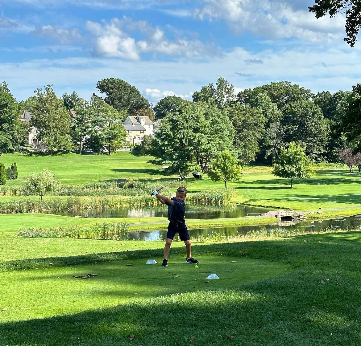 Person playing golf on a sunny green course near a pond, with trees and blue sky in the background