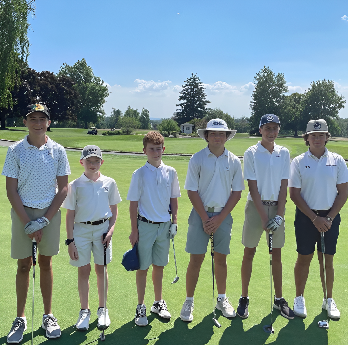 Group of young golfers on a green, holding clubs, wearing golf attire, posing outdoors on a sunny day.