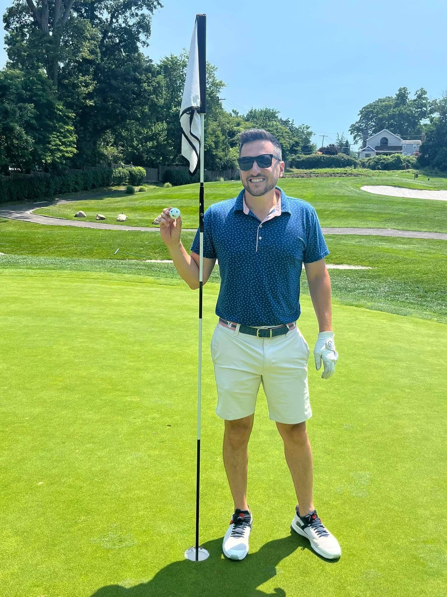 Man holding golf ball near flagstick on a green; sunny day.