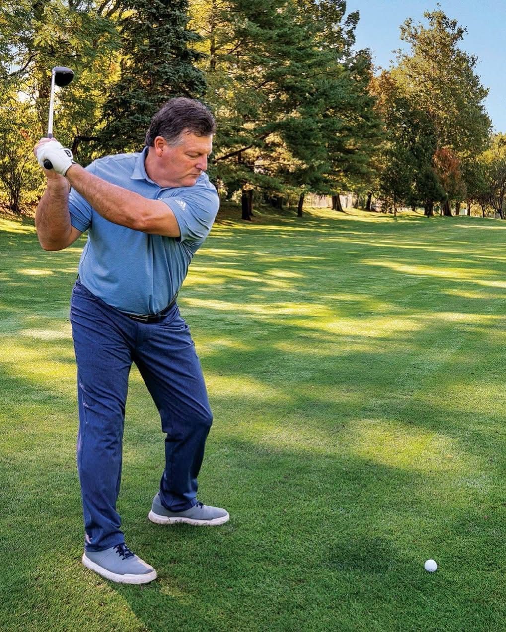 Man swinging a golf club on a green course; wearing blue shirt and pants.