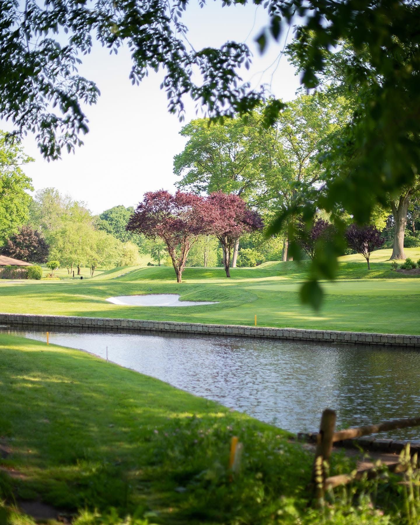 Green golf course with water feature, lush trees, and a cloudy sky.